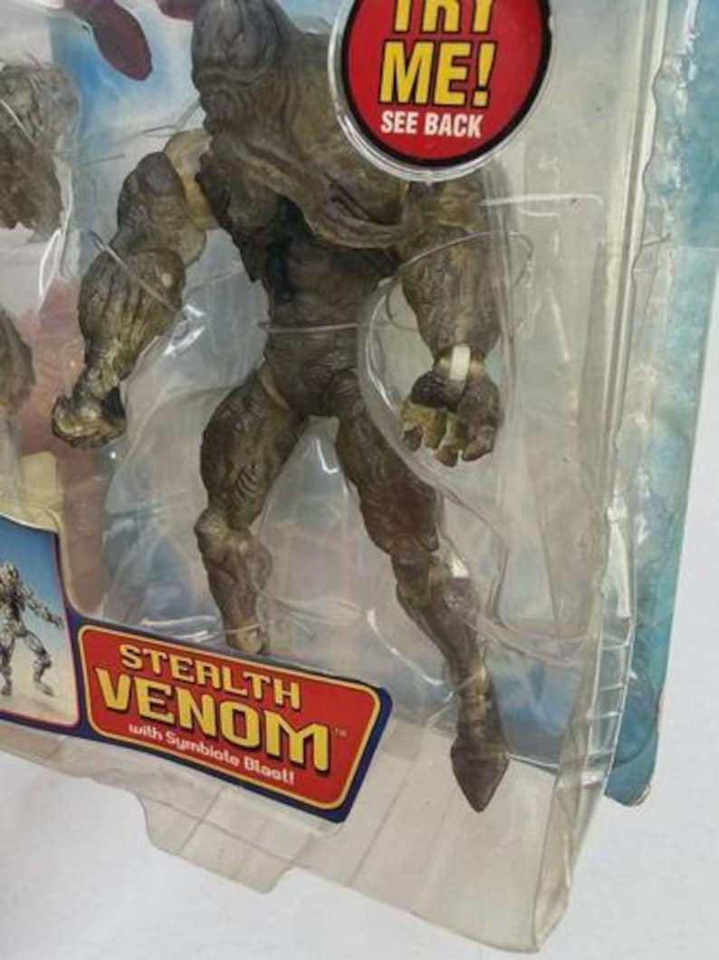 May include: Stealth Venom action figure in a clear plastic package. The figure is primarily olive green and brown, with detailed musculature. The packaging includes the text "TRY ME! SEE BACK" and "STEALTH VENOM with Symbiote Blast!"