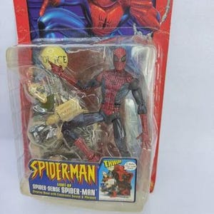 Peut inclure: Figurine de Spider-Man dans un emballage plastique. La figurine est rouge et noire, avec un costume d&eacute;taill&eacute;. L'emballage pr&eacute;sente le logo Spider-Man et les mots "Spider-Sense Spider-Man". L'arri&egrave;re-plan montre une illustration de bande dessin&eacute;e de Spider-Man.