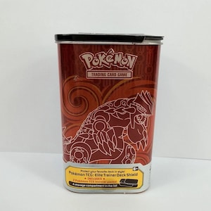 May include: A red Pokémon Trading Card Game storage tin featuring a Groudon illustration. The front displays the Pokémon logo, text reading Pokémon TCG: Elite Trainer Deck Shield, and details about two included booster packs and a lid compartment.