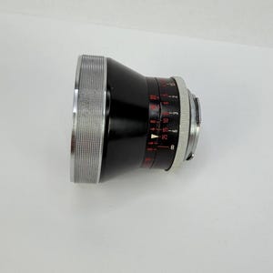 Puede incluir: Un objetivo de c&aacute;mara negro con detalles plateados. El objetivo tiene un anillo plateado estriado y marcas rojas y blancas de apertura. Dise&ntilde;ado para fotograf&iacute;a.