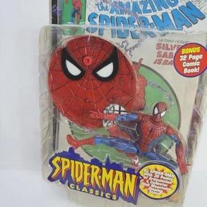 May include: Spider-Man action figure in a plastic package. The figure is red and blue, with a Spider-Man mask and web design. The packaging includes the text "SPIDER-MAN CLASSICS" and "the AMAZING SPIDER-MAN". A bonus 32-page comic book is included.