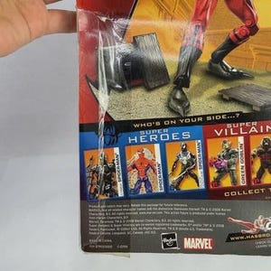 May include: A red and black superhero action figure in a cardboard box. The packaging shows other heroes and villains, with the Marvel logo. Text includes "Who's on your side...?" and "even SPIDER-MAN!" on the packaging.