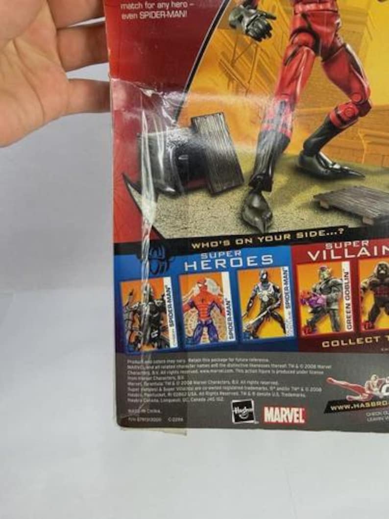 May include: A red and black superhero action figure in a cardboard box. The packaging shows other heroes and villains, with the Marvel logo. Text includes "Who's on your side...?" and "even SPIDER-MAN!" on the packaging.