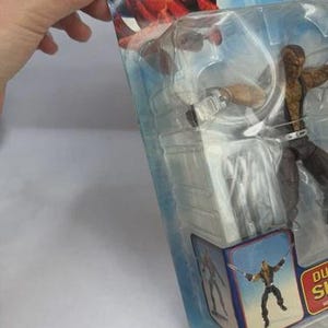 May include: An action figure of a character from The Amazing Spider-Man series, packaged in a clear plastic blister pack. The figure is brown and black, with a belt and a weapon. The packaging features Spider-Man graphics and the character's name.