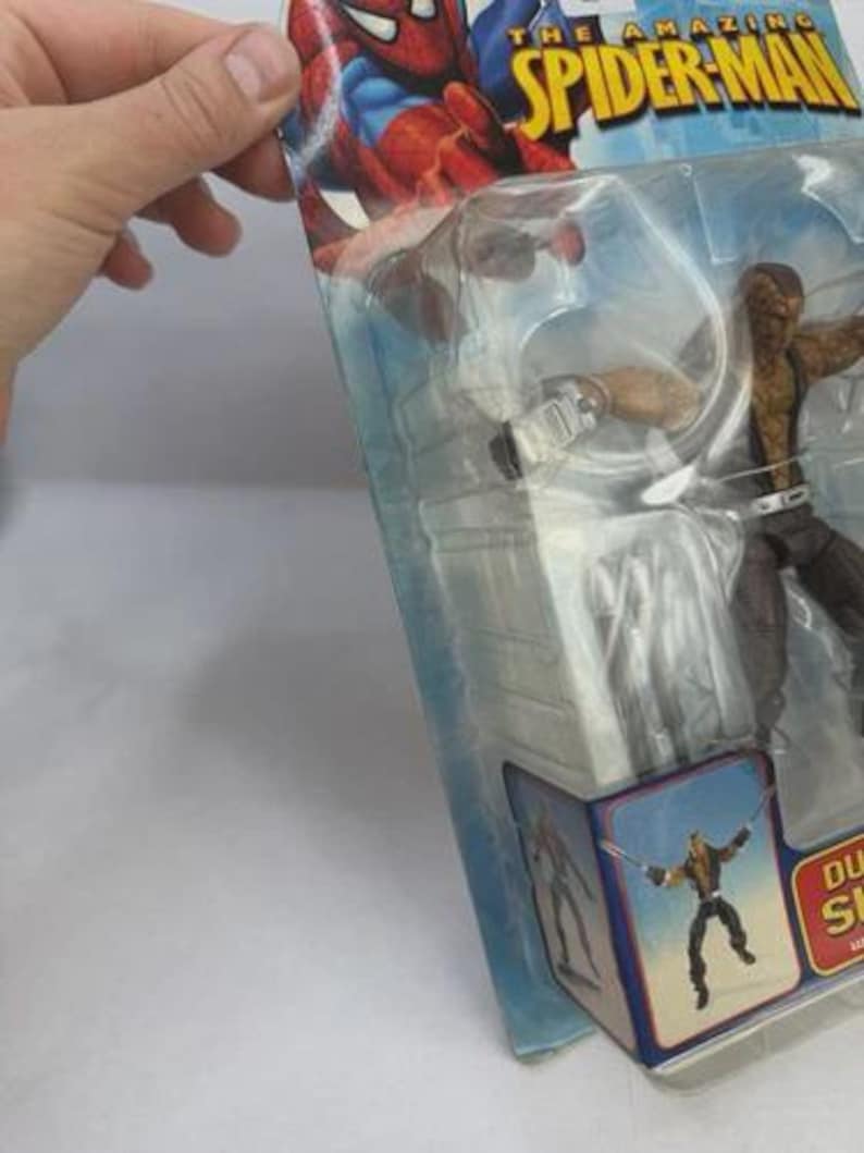 May include: An action figure of a character from The Amazing Spider-Man series, packaged in a clear plastic blister pack. The figure is brown and black, with a belt and a weapon. The packaging features Spider-Man graphics and the character's name.