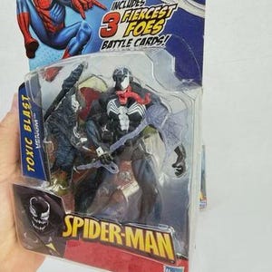 May include: A Spider-Man action figure toy package featuring the Venom character. The package includes the text "Toxic Blast Venom" and "Spider-Man". The packaging also includes a Spider-Man graphic and the text "Includes 3 Fiercest Foes Battle Cards!"