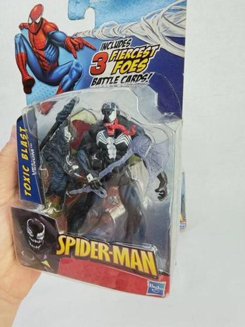 May include: A Spider-Man action figure toy package featuring the Venom character. The package includes the text "Toxic Blast Venom" and "Spider-Man". The packaging also includes a Spider-Man graphic and the text "Includes 3 Fiercest Foes Battle Cards!"