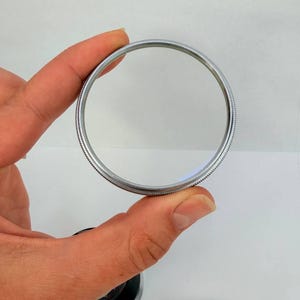 Puede incluir: Un filtro de lente de c&aacute;mara circular y transparente con un borde de metal plateado. El filtro se sostiene entre los dedos sobre un fondo blanco. Las palabras "ZEISS IKON" son visibles en un anillo plateado separado.