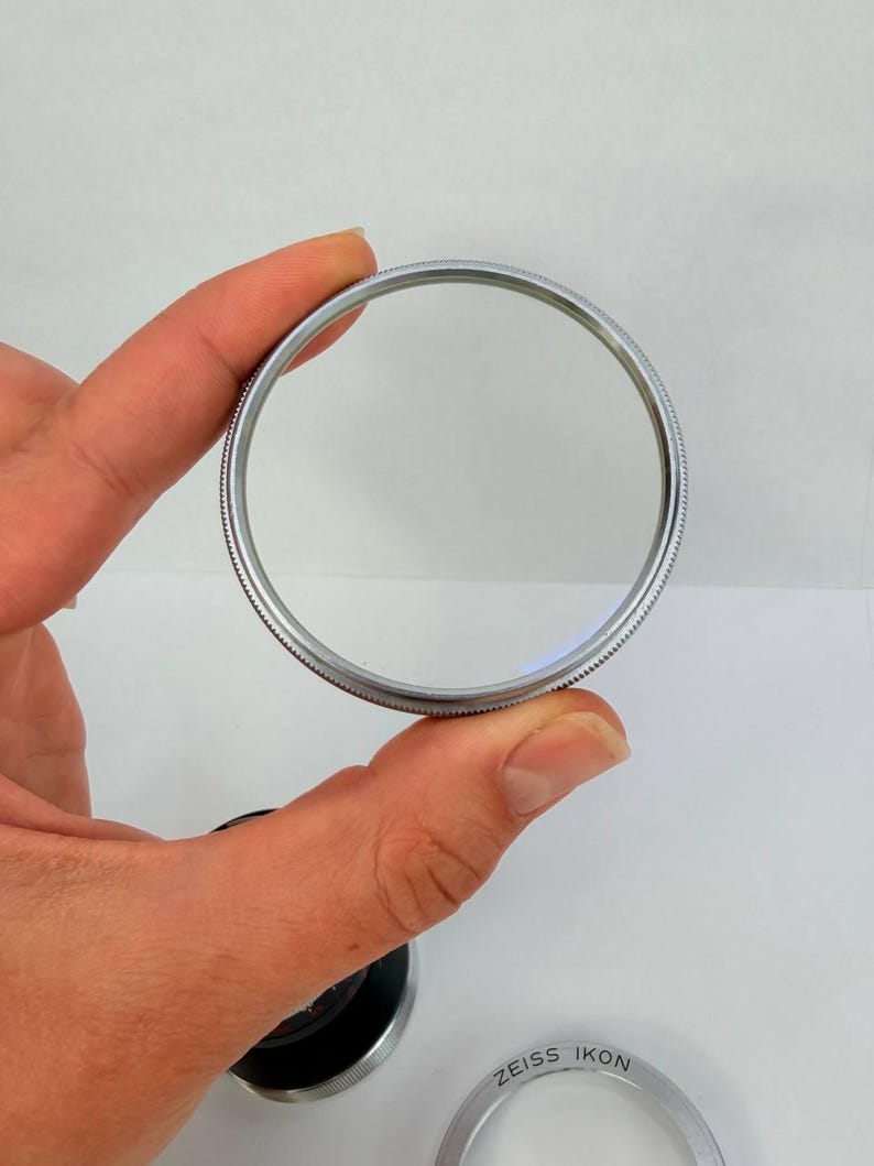 Puede incluir: Un filtro de lente de c&aacute;mara circular y transparente con un borde de metal plateado. El filtro se sostiene entre los dedos sobre un fondo blanco. Las palabras "ZEISS IKON" son visibles en un anillo plateado separado.