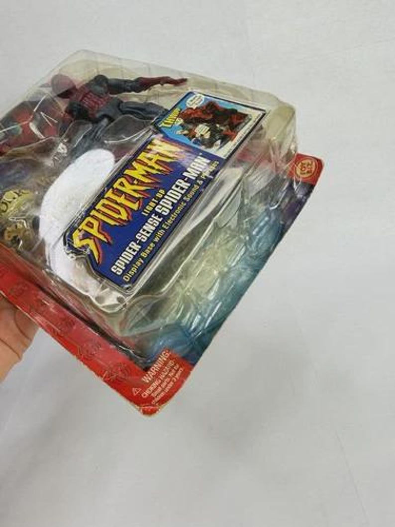 Peut inclure: Une figurine de Spider-Man dans un emballage en plastique. L'emballage comprend le texte "Spider-Man" et "Spider-Sense Spider-Man". La figurine est rouge et noire. L'emballage a une palette de couleurs rouge et bleu.