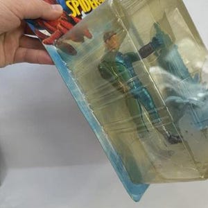 May include: Action figure of a character from The Amazing Spider-Man, in a sealed plastic package. The figure is wearing a green and blue costume. The packaging features the Spider-Man logo and artwork.