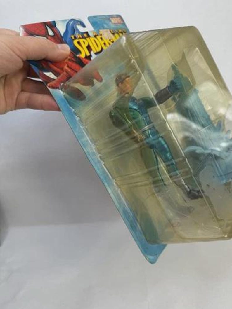 May include: Action figure of a character from The Amazing Spider-Man, in a sealed plastic package. The figure is wearing a green and blue costume. The packaging features the Spider-Man logo and artwork.