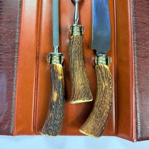 May include: A vintage carving set with three pieces: a sharpening steel, a carving fork, and a carving knife. The handles are made of textured, brown and tan antler material. The set is displayed on a brown leather-like surface.