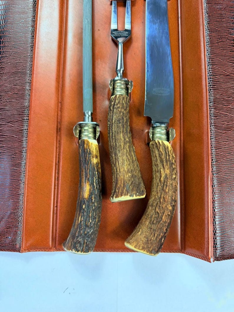 May include: A vintage carving set with three pieces: a sharpening steel, a carving fork, and a carving knife. The handles are made of textured, brown and tan antler material. The set is displayed on a brown leather-like surface.