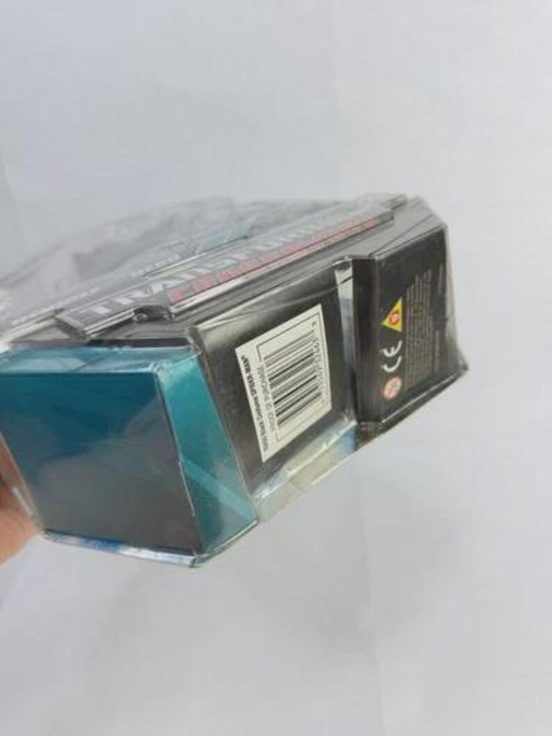 Puede incluir: Un producto empaquetado con una caja negra y turquesa, envuelto en pl&aacute;stico transparente. La caja tiene un c&oacute;digo de barras y texto. El producto es parcialmente visible, con texto rojo y blanco en la parte superior.