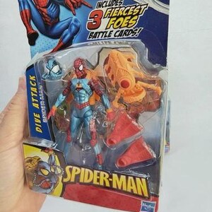 May include: Spider-Man action figure in a plastic package. The figure is blue, red, and orange. The packaging includes the text "DIVE ATTACK" and "SPIDER-MAN". The package also includes the text "INCLUDES 3 FIERCEST FOES BATTLE CARDS!"