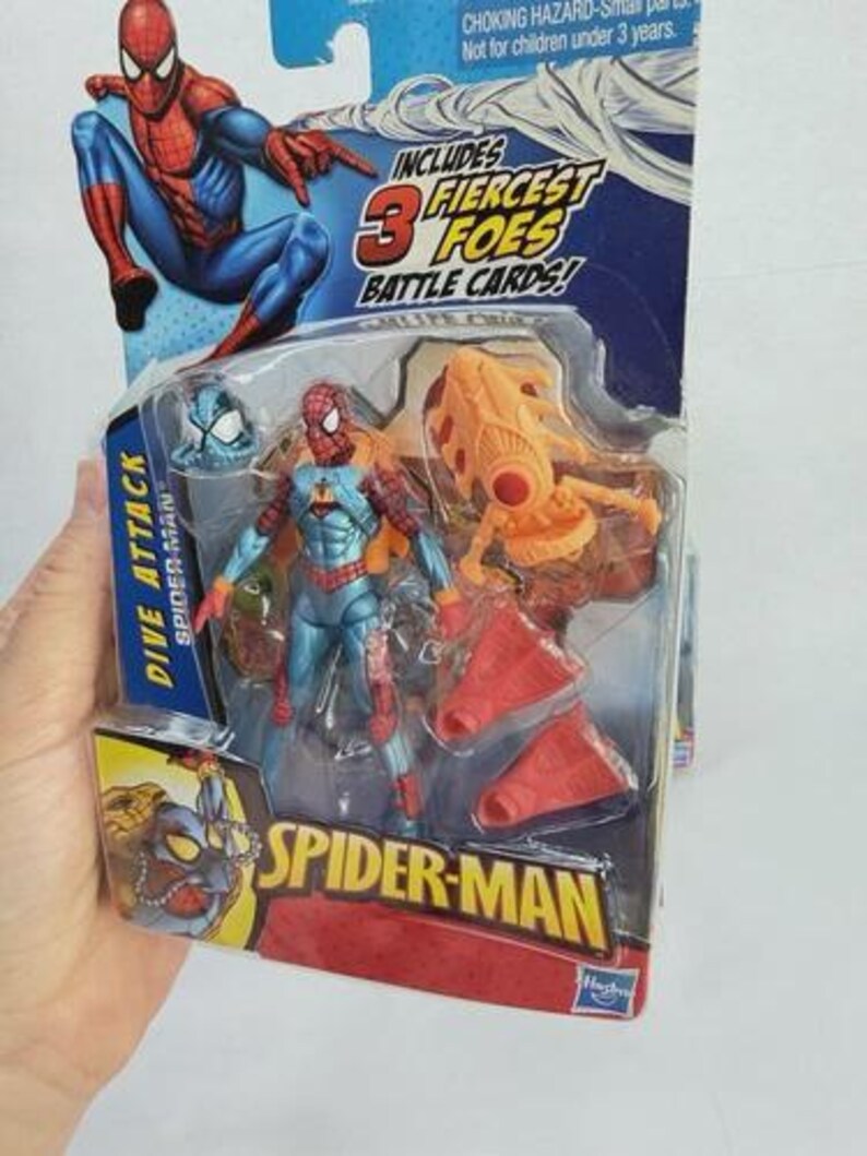 May include: Spider-Man action figure in a plastic package. The figure is blue, red, and orange. The packaging includes the text "DIVE ATTACK" and "SPIDER-MAN". The package also includes the text "INCLUDES 3 FIERCEST FOES BATTLE CARDS!"