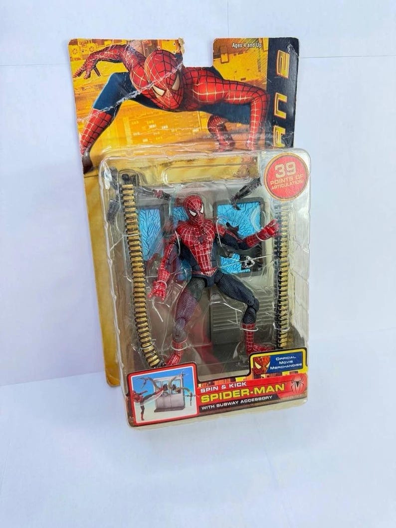 May include: Spider-Man action figure in a clear plastic package. The figure is red and black, with multiple points of articulation. The packaging includes the text "SPIN & KICK SPIDER-MAN WITH SUBWAY ACCESSORY" and "39 POINTS OF ARTICULATION".