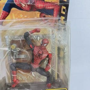 May include: A Spider-Man action figure in a clear plastic package. The figure is red and black, with a web pattern on the torso. The packaging includes the text "SHOOT AND SLIDE SPIDER-MAN" and "OFFICIAL Movie Merchandise".