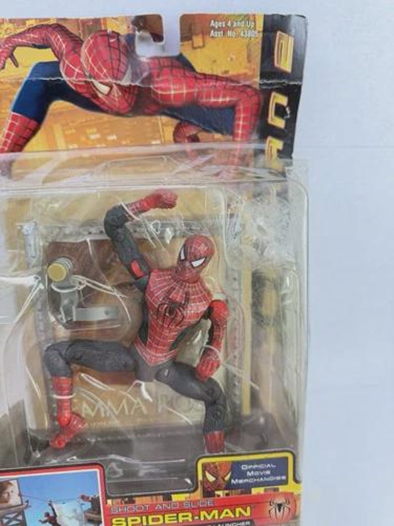 May include: A Spider-Man action figure in a clear plastic package. The figure is red and black, with a web pattern on the torso. The packaging includes the text "SHOOT AND SLIDE SPIDER-MAN" and "OFFICIAL Movie Merchandise".