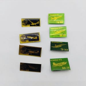 Pode incluir: Uma coleção de alfinetes de avião vintage em várias cores. Os alfinetes apresentam desenhos de aviões dourados e verdes com texto, incluindo "TU-104", "AK-40", "TU-144" e "IA-14". Alguns alfinetes têm um esquema de cores preto e dourado.