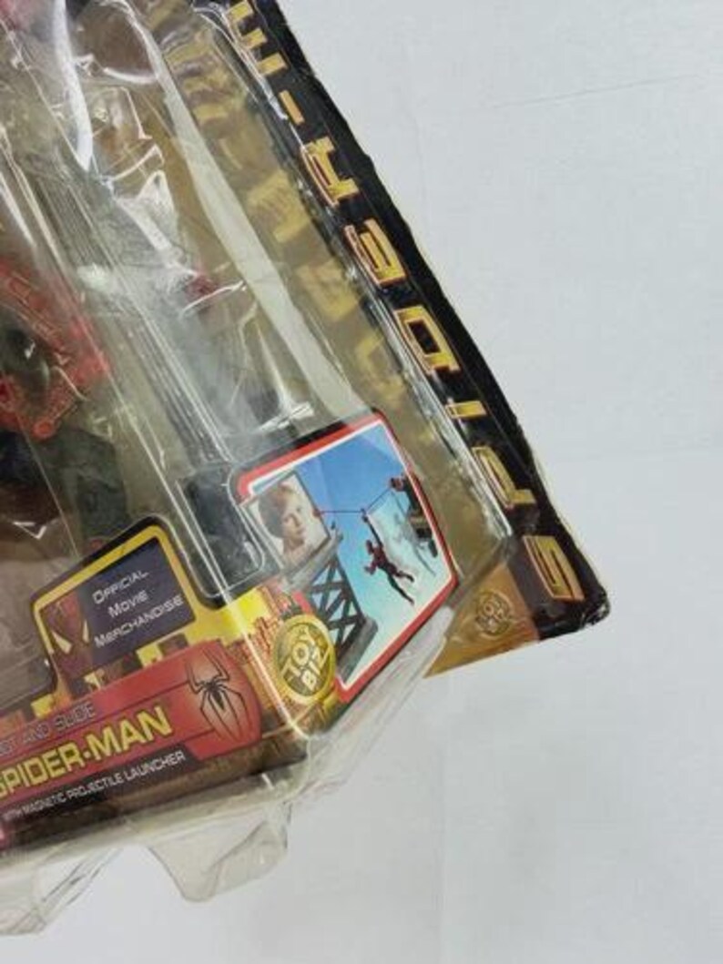 May include: Sealed Spider-Man toy package. The packaging is red and gold with the words "Spider-Man" and "Official Movie Merchandise." A small image shows Spider-Man swinging. The package includes a magnetic projectile launcher.