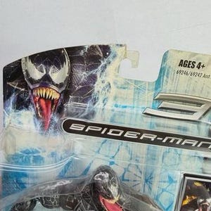 May include: A Spider-Man action figure of Venom, a black and white character with a long red tongue, is displayed in its original packaging. The packaging features the Venom character's face and the words "Spider-Man". The toy is recommended for ages 4+.
