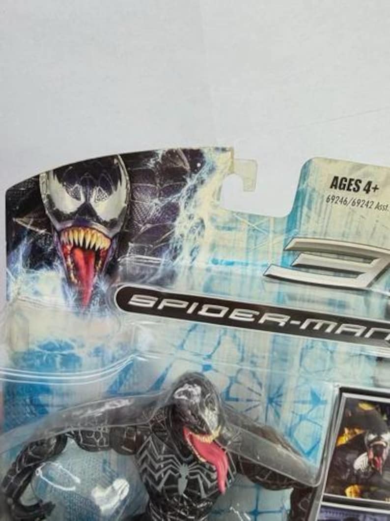May include: A Spider-Man action figure of Venom, a black and white character with a long red tongue, is displayed in its original packaging. The packaging features the Venom character's face and the words "Spider-Man". The toy is recommended for ages 4+.