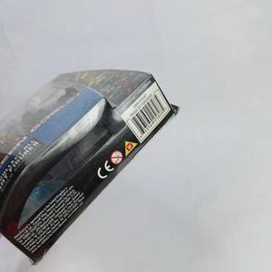 May include: A partially visible action figure package. The package is black with blue and red accents. A barcode and the CE mark are visible. The package is angled, with the top left corner closest to the viewer.