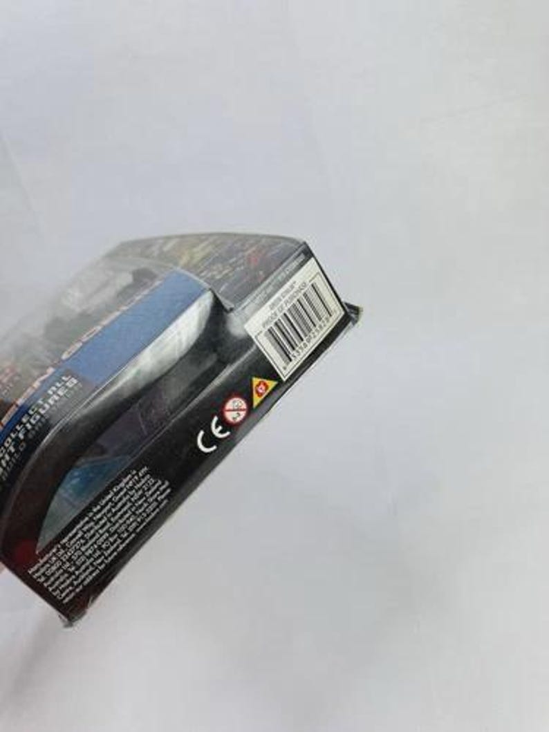 May include: A partially visible action figure package. The package is black with blue and red accents. A barcode and the CE mark are visible. The package is angled, with the top left corner closest to the viewer.