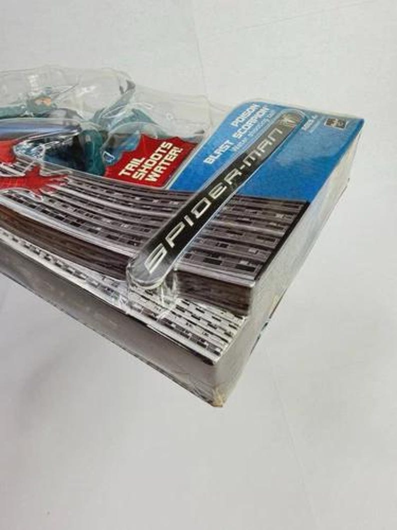 Puede incluir: Paquete sellado de figura de acci&oacute;n de Spider-Man. La caja es azul y presenta las palabras "Spider-Man" y "Poison Blast Scorpion". El paquete est&aacute; envuelto en pl&aacute;stico transparente.