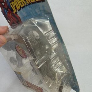 May include: Sealed action figure of Spider-Man in a clear plastic package. The packaging features the words "THE AMAZING SPIDER-MAN" in yellow and blue. The figure is partially visible, with a red and blue color scheme.