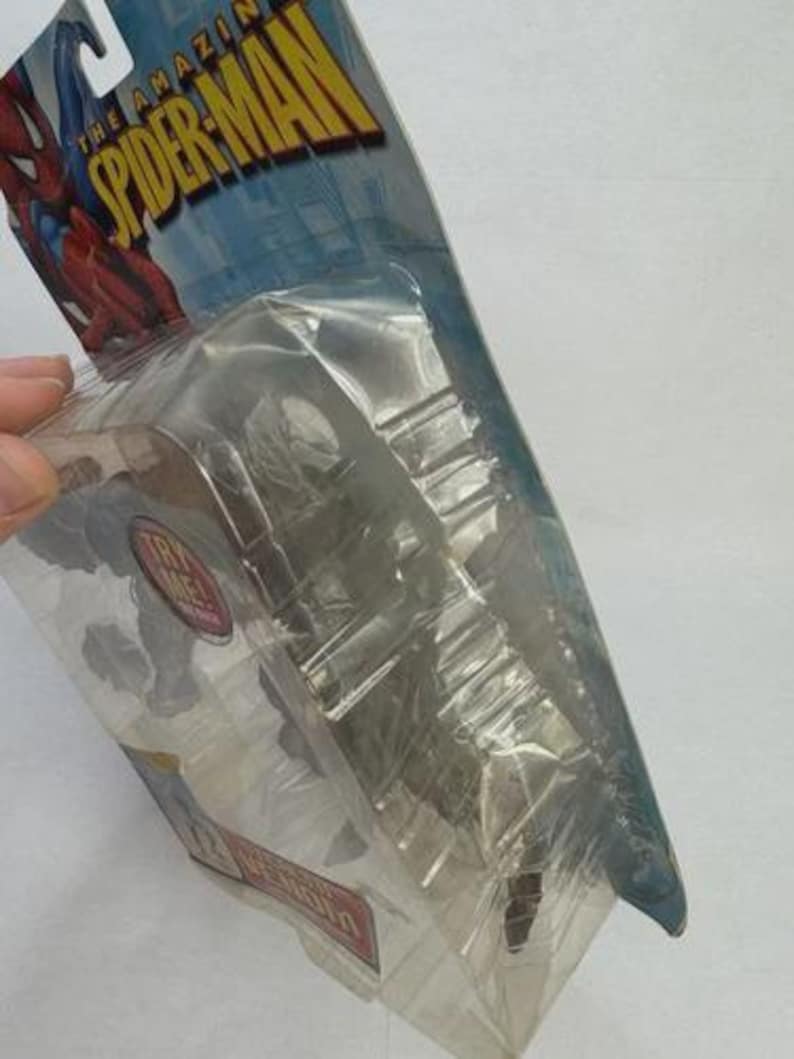 May include: Sealed action figure of Spider-Man in a clear plastic package. The packaging features the words "THE AMAZING SPIDER-MAN" in yellow and blue. The figure is partially visible, with a red and blue color scheme.