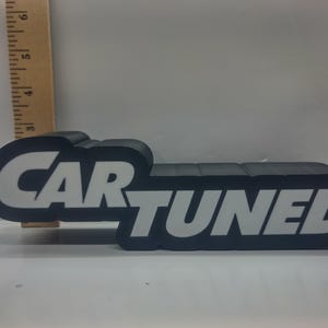May include: Black and white "CAR TUNED" sign. The sign is a rectangular shape with rounded edges. The text is in a bold, sans-serif font. A wooden ruler is in the background.