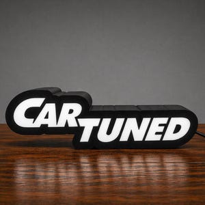 May include: A black and white LED sign with the words "CAR TUNED" in a bold, stylized font. The sign is rectangular with rounded edges and sits on a wooden surface. A black power cord extends from the sign.