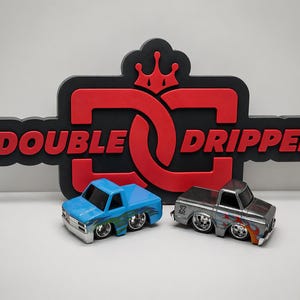 May include: Two toy pickup trucks, one blue and one silver, are displayed in front of a black and red sign that reads "DOUBLE DRIPPED" with a crown graphic. The blue truck has a flame design, and the silver truck has a "Z" logo.