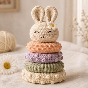 May include: A crochet stacking toy featuring a beige bunny head topper with a white flower accent. The toy consists of four stacked rings in peach, lavender, sage green, and cream, each with a textured bobble or ribbed stitch pattern.