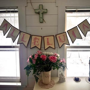 May include: A banner with the word "ALLELUIA" in pink letters hangs above a vase of pink roses and white flowers. A statue of the Virgin Mary stands on a windowsill, with a green cross on the wall.