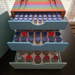 May include: A multi-colored, rainbow-striped storage box with three light blue drawers. Each drawer contains multiple small glass vials with blue and red caps. The box has a handle for easy carrying.