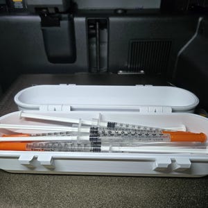 May include: A white, oval-shaped case containing several syringes with orange caps. The syringes are clear with black measurement markings. The case is open, revealing the contents inside. The background is a dark interior.