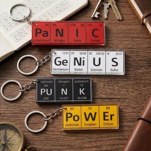 May include: Four keychains with periodic table designs spelling out "PANIC", "GENIUS", "PUNK", and "POWER". Each keychain has a silver chain and ring. The keychains are rectangular with a clear, glossy finish. The background is a wooden surface.