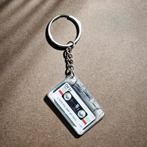 May include: A clear acrylic keychain shaped like a cassette tape. The tape has a white label with the text "TAPE-UP AND TEA BOY" and "FE 90". The keychain is attached to a silver chain and ring.