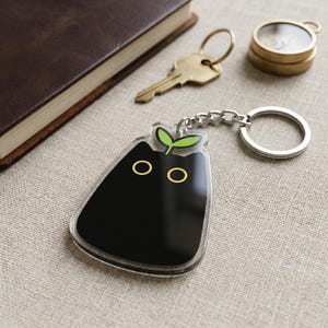 May include: A clear acrylic keychain featuring a black cat design with yellow eyes and a green sprout on top. The keychain is attached to a silver chain and ring. Other items in the background include a brown notebook, a key, and a compass.