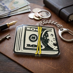 May include: A rectangular keychain featuring a stack of money with a yellow band. The keychain has a silver chain and clasp. Other items in the image include a pen, key, coins, and a leather-bound notebook.