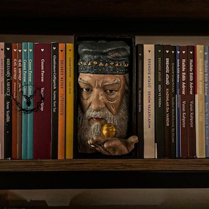 Könnte beinhalten: Ein Bücherregal mit einer geschnitzten Holzskulptur eines alten Mannes mit langem Bart, der eine goldene Kugel hält. Die Skulptur befindet sich zwischen einer Reihe farbiger Bücher mit türkischen Titeln.