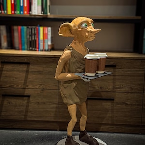 Puede incluir: Estatua detallada de un personaje ficticio, sosteniendo una bandeja con dos tazas de café. La figura es de color marrón claro con orejas grandes y ojos verdes, vistiendo una túnica y zapatos marrones. La estatua se encuentra sobre una base redonda gris.
