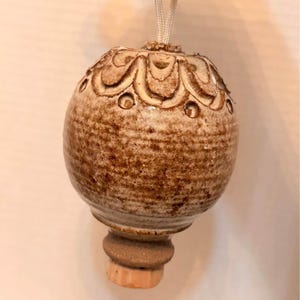 May include: A round, speckled brown ceramic ornament with a textured surface featuring a carved scalloped pattern near the top. The ornament has a light-coloured cord attached to the top for hanging and a small, unglazed clay base at the bottom.