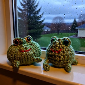 May include: Two handmade green crochet frog toys with large black eyes and red smiles sit on a windowsill. The frogs have a textured appearance and are positioned in front of a window with a rainy outdoor scene.