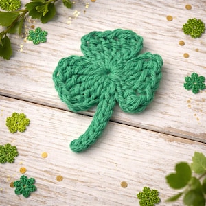 Può includere: Un trifoglio verde all'uncinetto fatto a mano, simbolo del giorno di San Patrizio, poggia su una superficie di legno bianco. Il trifoglio è circondato da piccole decorazioni di trifogli verdi glitterati e coriandoli dorati. Il trifoglio all'uncinetto è un trifoglio a tre foglie.