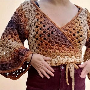 May include: A handmade crochet wrap top in shades of brown, tan, and purple. The top has a V-neck, long bell sleeves, and a tie waist. The openwork design adds a delicate touch to this cosy garment.
